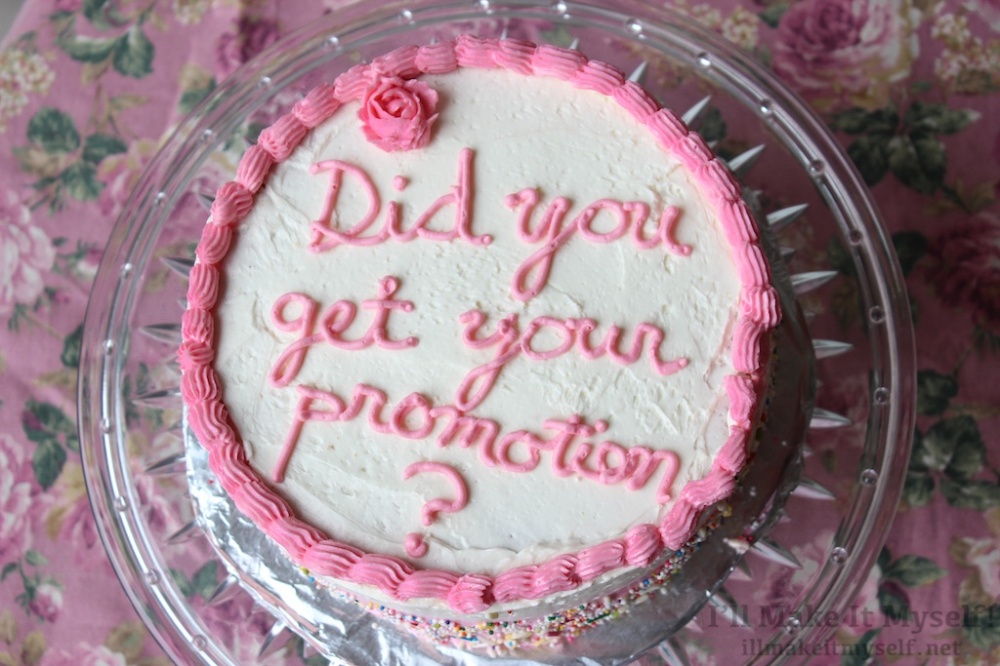 A three-layer round cake with pink and white frosting. There is a border piped around the top. The text reads, “Did you get your promotion?” The sides of the cake are white to pink ombré and covered in sprinkles. 