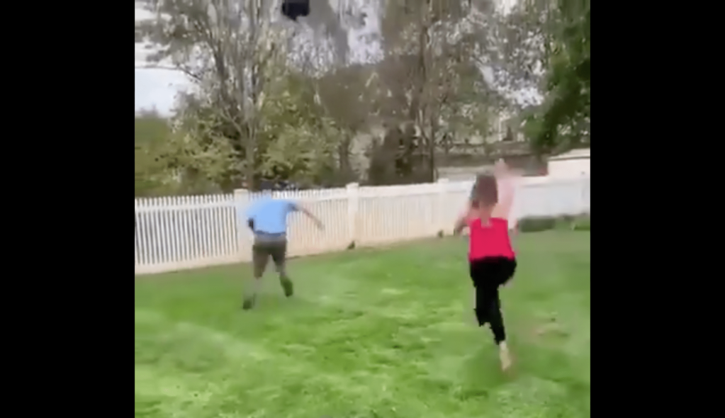 The father-to-be and a party-goer chase after the escaped black balloon towards a white picket fence.